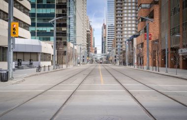 A shot down a street in Calgary's Downtown West, with LRT tracks running through the street. Weather is partly cloudy.