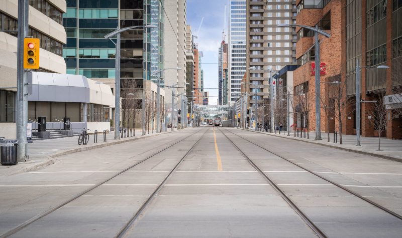 A shot down a street in Calgary's Downtown West, with LRT tracks running through the street. Weather is partly cloudy.