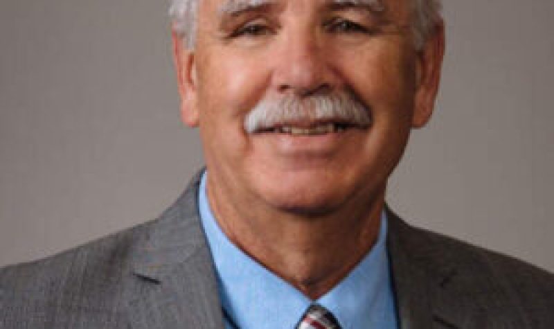 Photo of Stan Peterson standing in front of a wall wearing a grey suit and blue shirt.