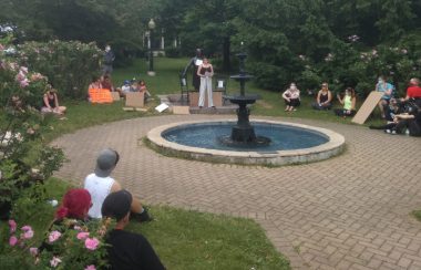 People sitting in the park and listening to Cheyanne Thorpe discuss the negative impacts of the Sir John A MacDonald statue, which stands behind her.