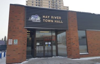 La facade d'un petit édifice brun, ou est inscrit «Hay River Town Hall», avec un immeuble en arrière plan .