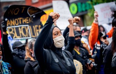 Une femme noire a le poing levé dans une manifestation avec des pancartes contre les actes de racismes.