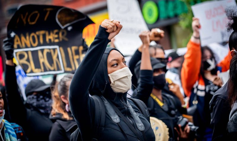 Une femme noire a le poing levé dans une manifestation avec des pancartes contre les actes de racismes.