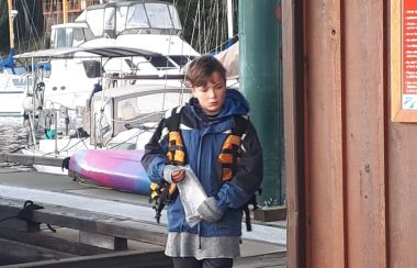 Youth in life jacket walks on dock with yacht in background