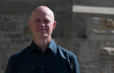 A headshot of Dominion Sculptor John-Philippe Smith, who is bald and wearing a dark collared shirt and standing in front of a stone wall.