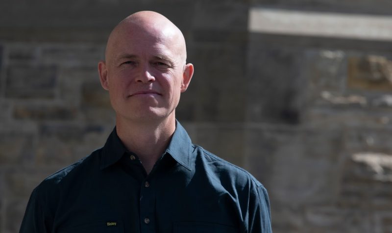 A headshot of Dominion Sculptor John-Philippe Smith, who is bald and wearing a dark collared shirt and standing in front of a stone wall.