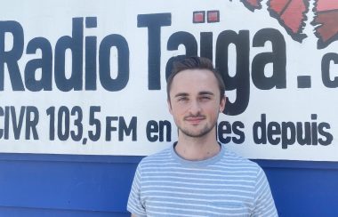 Damian Benoit vêtu d'un chandail rayé, devant une pancarte du logo de radio Taïga.