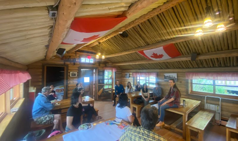 Un groupe est assis aux tables dans un cabane de bois rond et écoutent la présentation d'une femme.