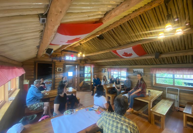 Un groupe est assis aux tables dans un cabane de bois rond et écoutent la présentation d'une femme.