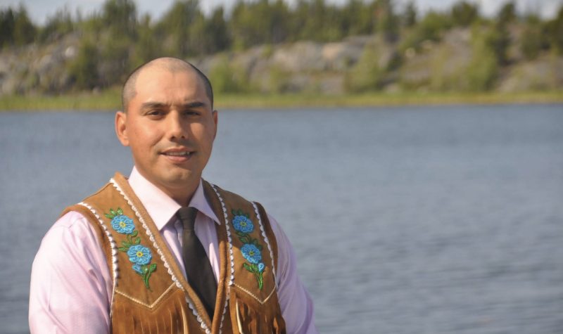 Un homme vêtu d'une veste traditionnelle aux du peuple Déné, pose devant un lac.