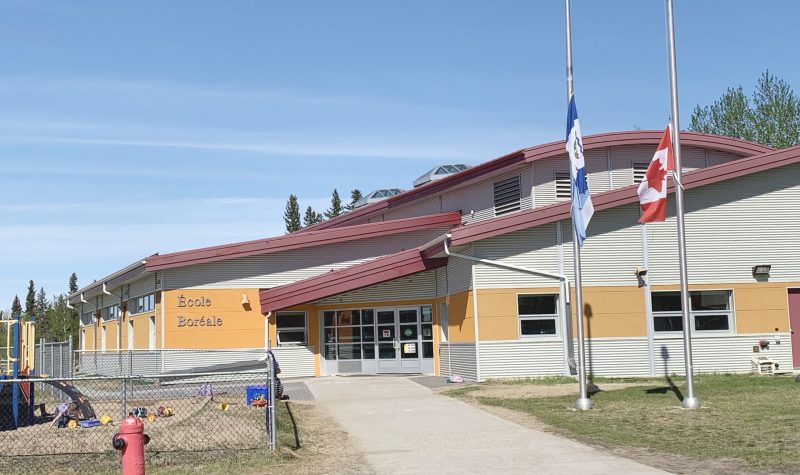 En été, la devanture d'une école. Les drapeaux du Canada et des Territoires du Nord-Ouest sont en berne.