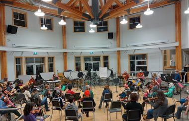 A view from the back of the Valley Youth Fiddlers in action at Smithers Secondary School in a large room with a high ceiling.