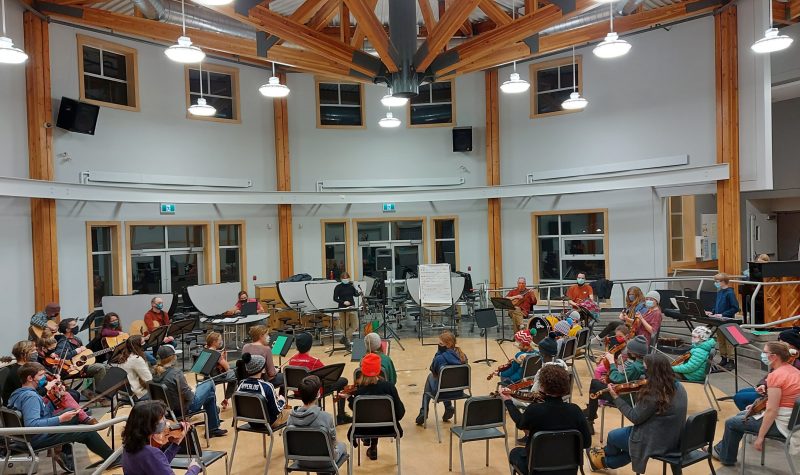 A view from the back of the Valley Youth Fiddlers in action at Smithers Secondary School in a large room with a high ceiling.