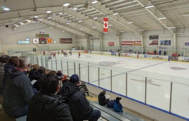 A view of the ice in a hockey arena, taken from the stands with people seated and looking towards the rink.