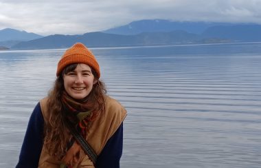 A young woman smiles at the camera with a background of ocean and mountains.