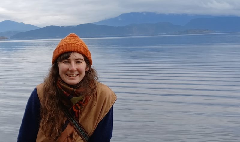 A young woman smiles at the camera with a background of ocean and mountains.