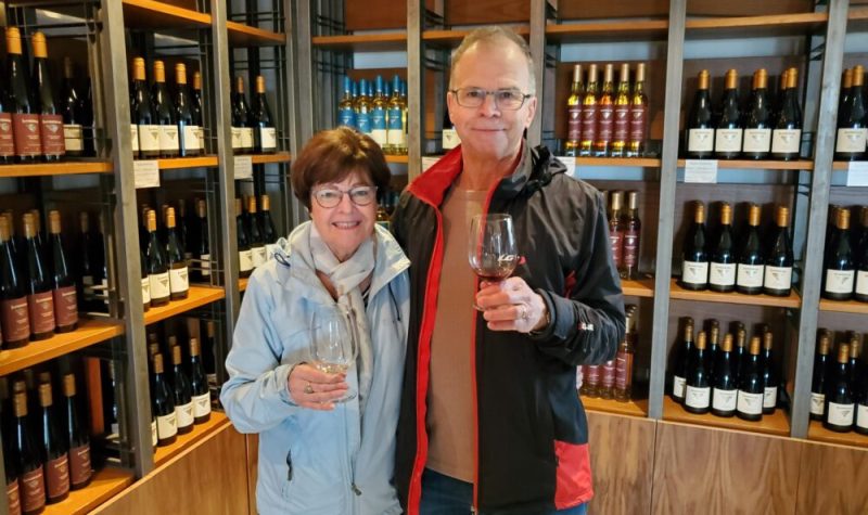 Un couple de sexagénaire tenant chacun un verre de vin devant un étagère de bouteilles de vin diversifiées.