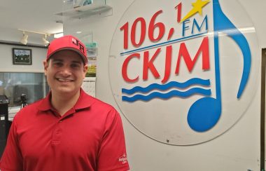 Un homme portant un chandail et une casquette rouge en avant du logo de Radio CKJM.