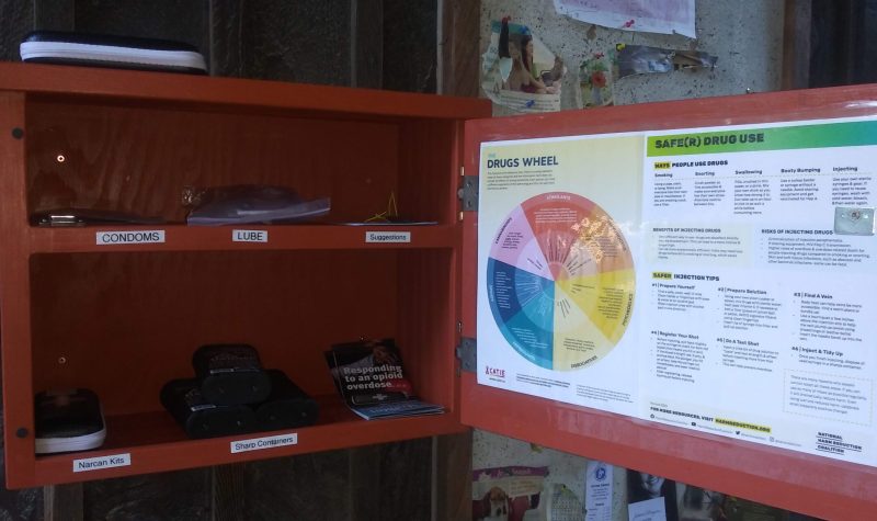 An orange box on a public wall, with the door ajar, revealing free naloxone kits, and condoms.