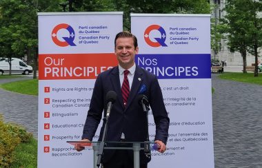Colin Standish standing at a podium with posters of the Canadian Party of Quebec's foundational principles in the background. The party's colours are orange and purple.