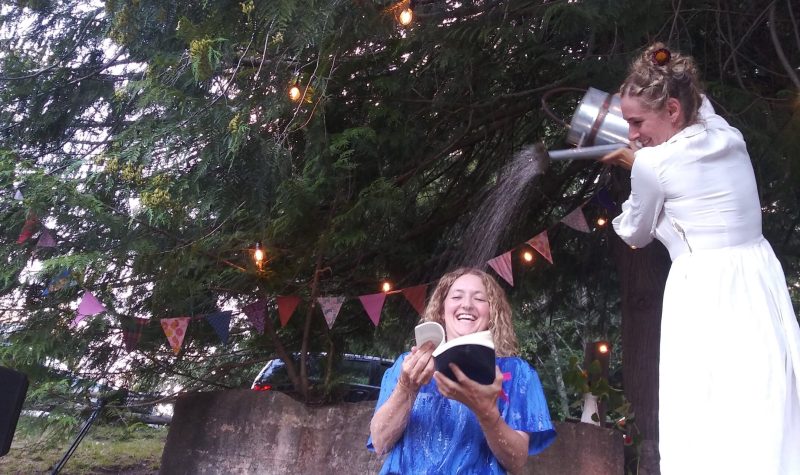 A woman in white pours water with a watering can onto a lady in blue, below her, holding a book.