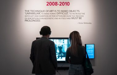 Two people standing at the Video poetry exhibit watching a screen with headphones on