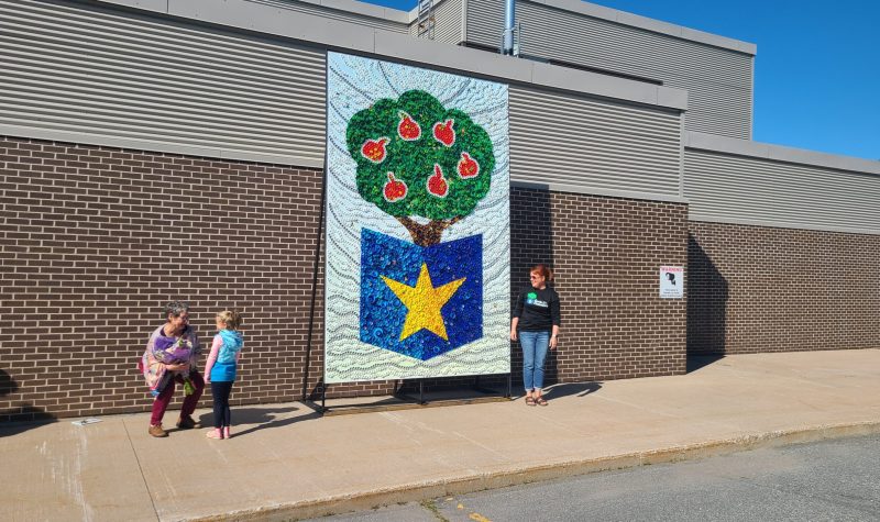 Au centre, la mosaique representant le logo de l'école, un pommier sur fond blanc sur un livre ouvert sur lequel on voit l'étoile jaune du drapeau Acadien. A gauche, l'artiste recoit un bouquet de fleurs de la part d'une élève, et a droite la directrice de l'école.