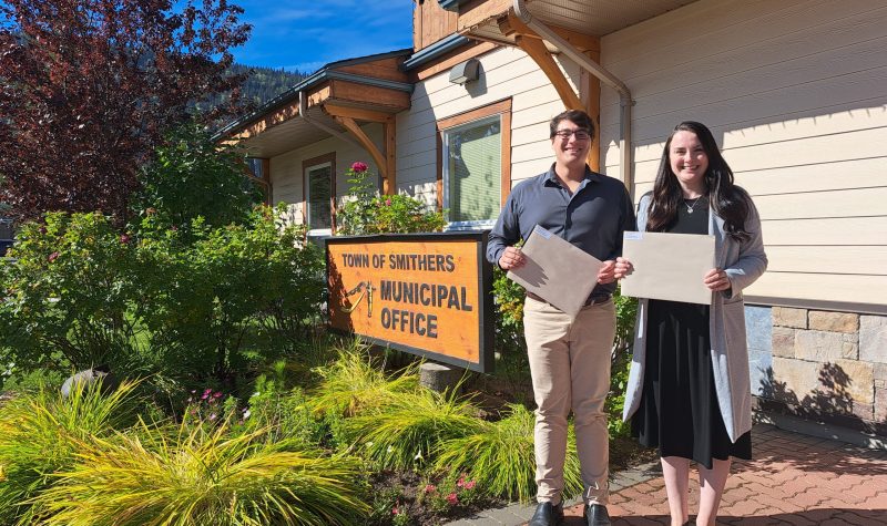 a man and a woman stand in front of the sign at the front of the Smithers town offices for council and mayor. They are each holding an envelope to represent mail-in voter registration packages.
