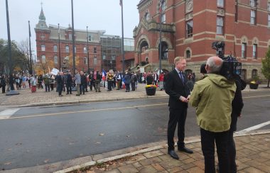 L'ancien ministre Dominic Cardy en costume sombre répond aux journalistes devant l'hôtel de ville ou se réunissent les premier manifestants.