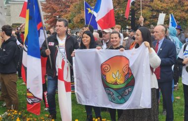 On vois des manifestants rassemblés lors du tintamarre du 25 octobre a Fredericton. Certains portent des drapeaux acadiens, d'autres ont des drapeaux autochtones.