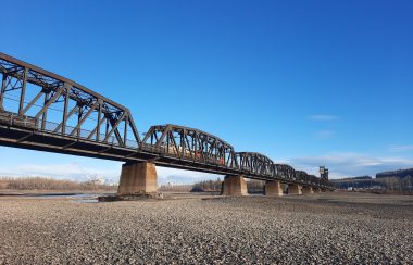 A train bridge which spans across the confluence of a dry river. The sky is blue with a couple of clouds low on the horizon.