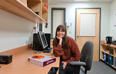 a woman sits at a long desk against a wall in a small room with a computer and photocopier. She is smiling and looking directly at the camera.