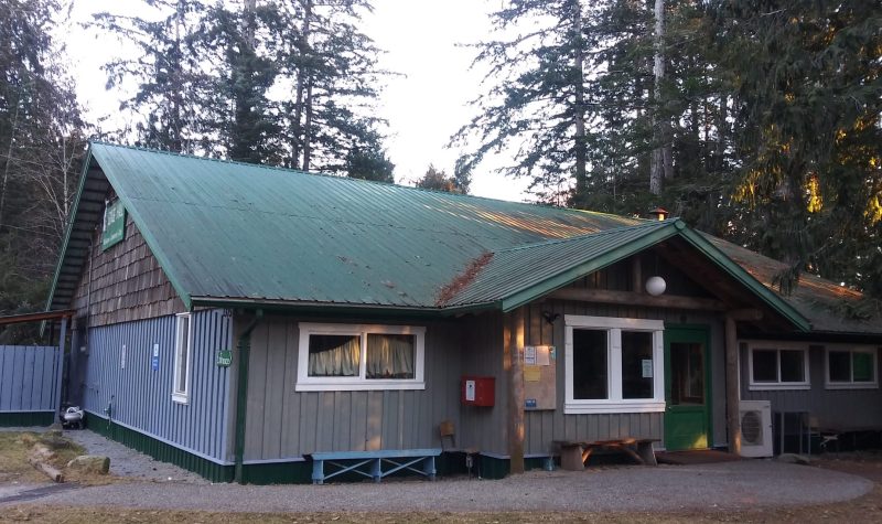 A large blue building sits idle under large cedar trees.