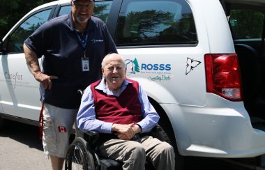 A man wearing a ROSSS hat and shirt stands in front of the ROSSS white mini van and behind an elderly man in a wheelchair, about to be loaded into the van. Photo courtesy of ROSSS.