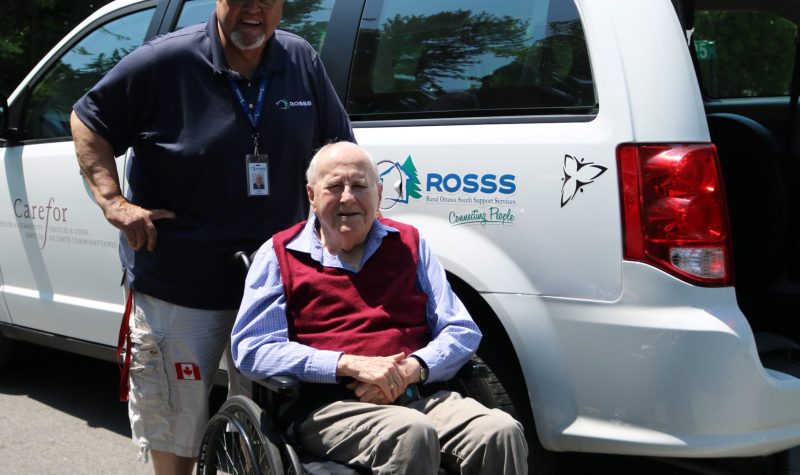 A man wearing a ROSSS hat and shirt stands in front of the ROSSS white mini van and behind an elderly man in a wheelchair, about to be loaded into the van. Photo courtesy of ROSSS.