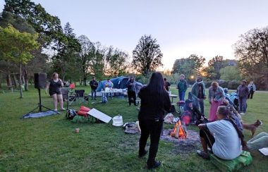 A group of folks in a park with a sunset in the background beyond trees. In the foreground the aforementioned folks with a group of them gathered around a fire. Tents are visible as is the sound system for the event.,