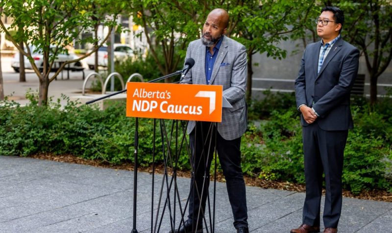 Deux hommes habillés en costard se tiennent derrière un podium. Sur le podium orange est inscrit « Alberta's NPD caucus ». Derrière eux, il y a des arbres et une rue avec quelques voitures.