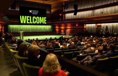 A crowd sitting in seats in an auditorium, facing a stage with a podium and a large, glowing 'WELCOME' sign with green letters. Above the word 'Welcome' is written 'Kingston Business Awards'.