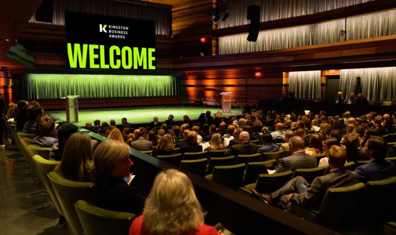 A crowd sitting in seats in an auditorium, facing a stage with a podium and a large, glowing 'WELCOME' sign with green letters. Above the word 'Welcome' is written 'Kingston Business Awards'.