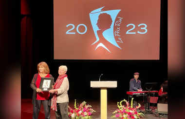 Claire Desrosiers (en chemise rouge) reçoit le Prix Riel des mains de son amie (à droite) qui porte une chemise blanche et le foulard du festival du voyageur. À l'arrière, un grand écran avec le logo du Prix Riel en blanc sur un fond orange.
