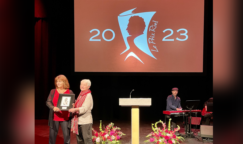 Claire Desrosiers (en chemise rouge) reçoit le Prix Riel des mains de son amie (à droite) qui porte une chemise blanche et le foulard du festival du voyageur. À l'arrière, un grand écran avec le logo du Prix Riel en blanc sur un fond orange.