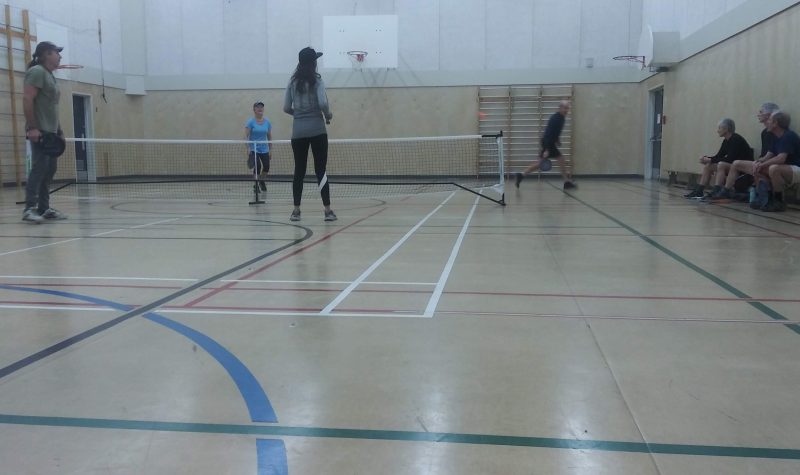 Four adults play pickleball in a school gym, as others watch.