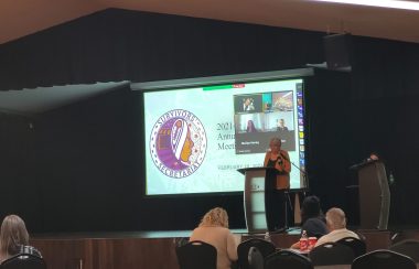 A woman stand's on a stage in front of people. A large projector screen is in the background.