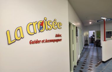 Le logo de la croisée, jaune sur mur blanc. A droite on apercois les bureaux .
