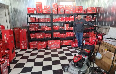 A man in a grey sweater stands in front of a shelf full of red Milwaukee equipment in boxes.