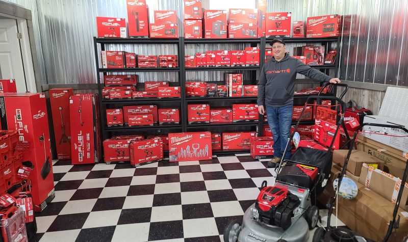 A man in a grey sweater stands in front of a shelf full of red Milwaukee equipment in boxes.
