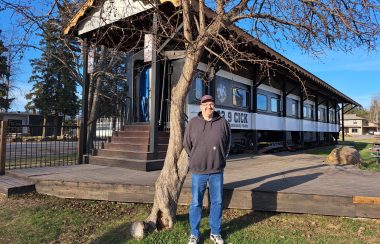 a man stands in front of the Smithers Community Radio radio station, which happens to be a 1929 CN Train car (on the tracks).