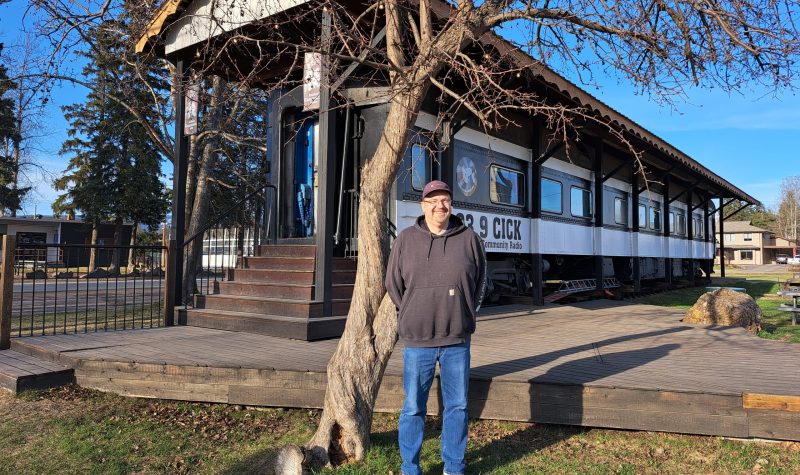 a man stands in front of the Smithers Community Radio radio station, which happens to be a 1929 CN Train car (on the tracks).