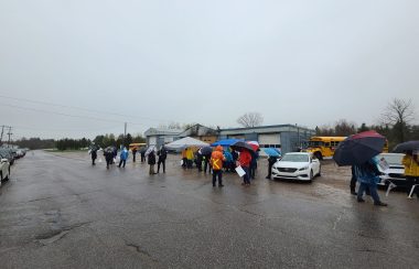 Union members wearing raincoats and carrying umbrellas and signs form a picket line outside a bus garage.
