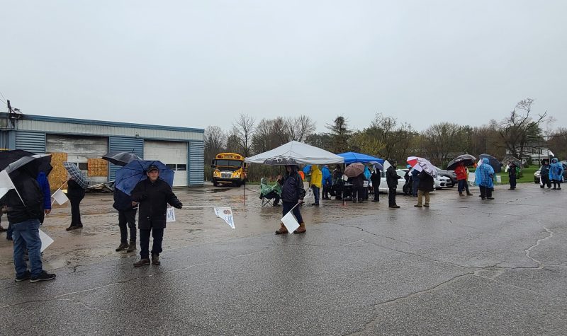 Bus drivers on strike picket in the rain outside of a blue garage.